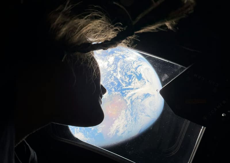 Silhouette of an Artemis II astronaut looking through the Orion spacecraft window at a glowing Earth from space.
